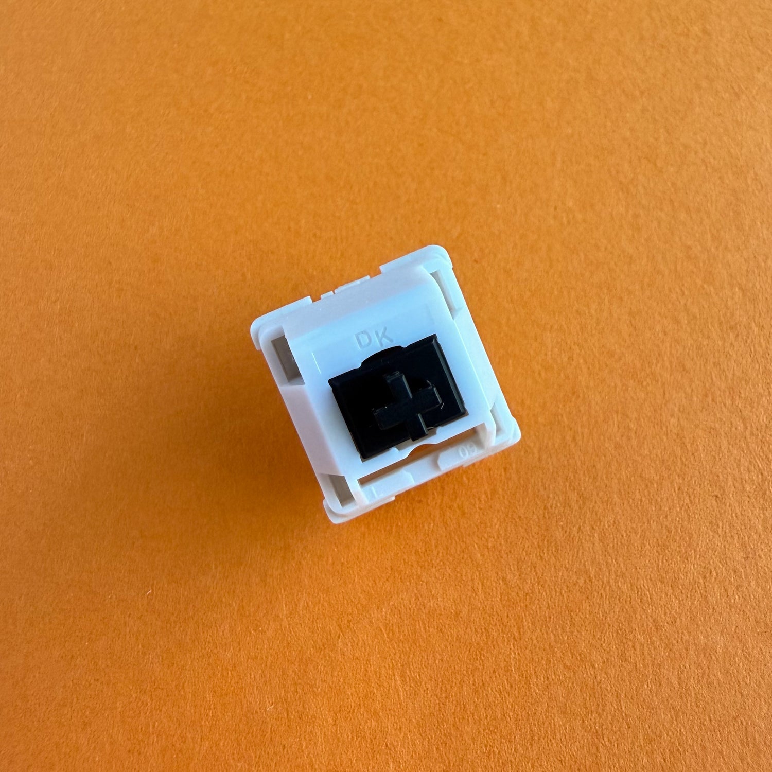 A tactile keyboard switch on an orange background. The switch component is black, and the casing is white with the letters "DK" imprinted into it.