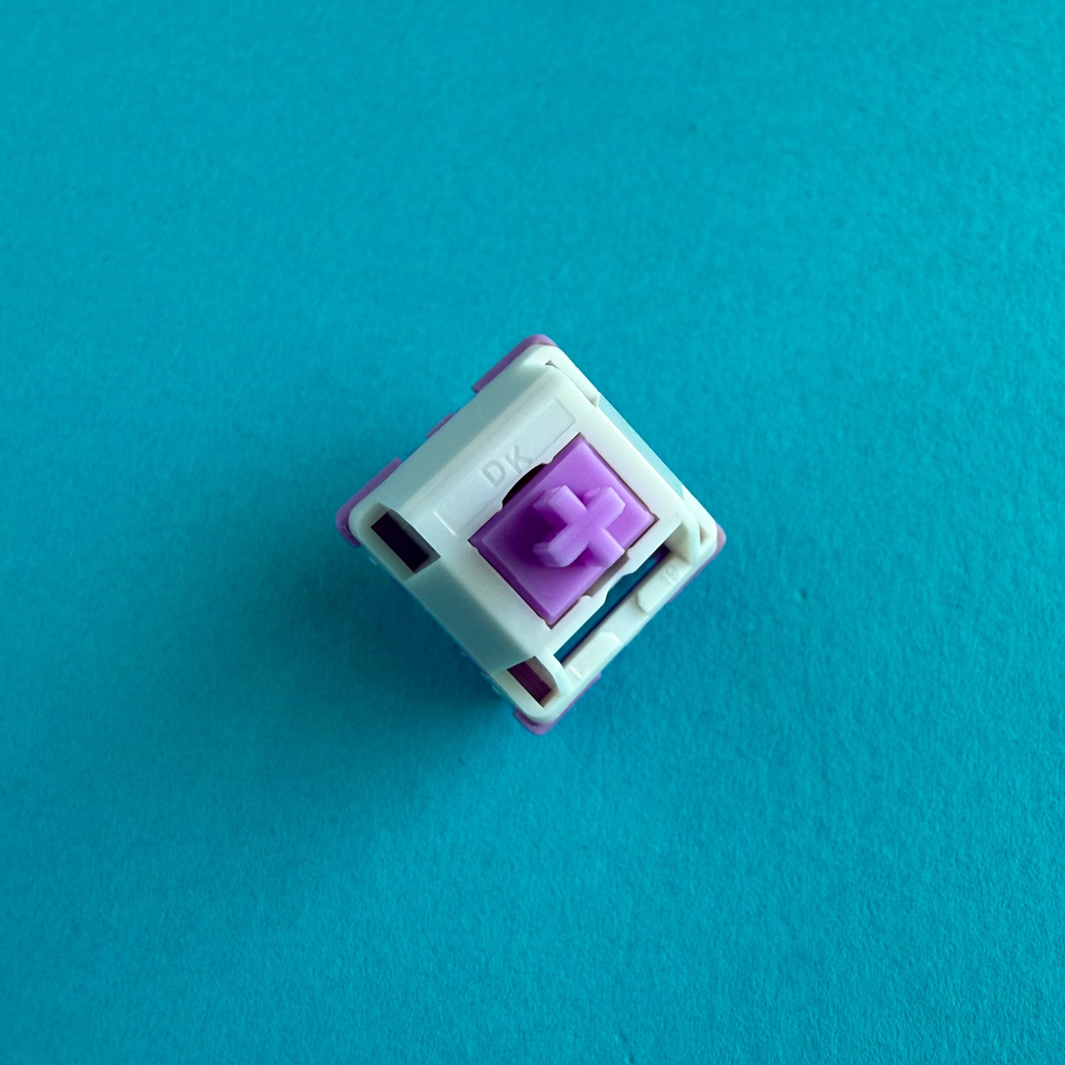 A linear keyboard switch on a blue background. The switch component is purple, the bottom of the casing is also purple, and the top of the casing is white with the letters "DK" imprinted into it.