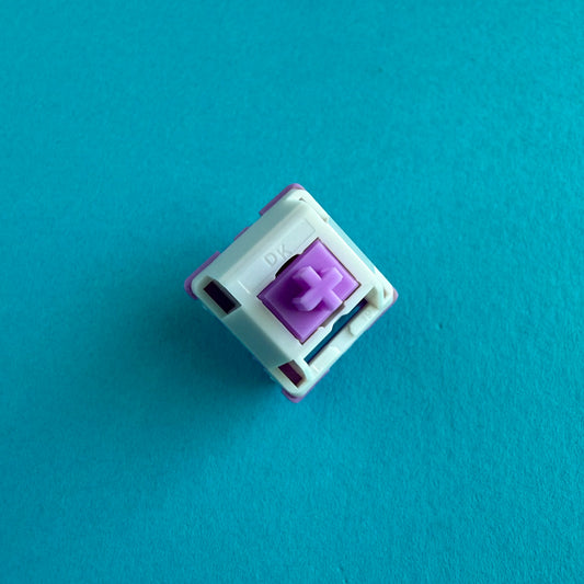 A linear keyboard switch on a blue background. The switch component is purple, the bottom of the casing is also purple, and the top of the casing is white with the letters "DK" imprinted into it.