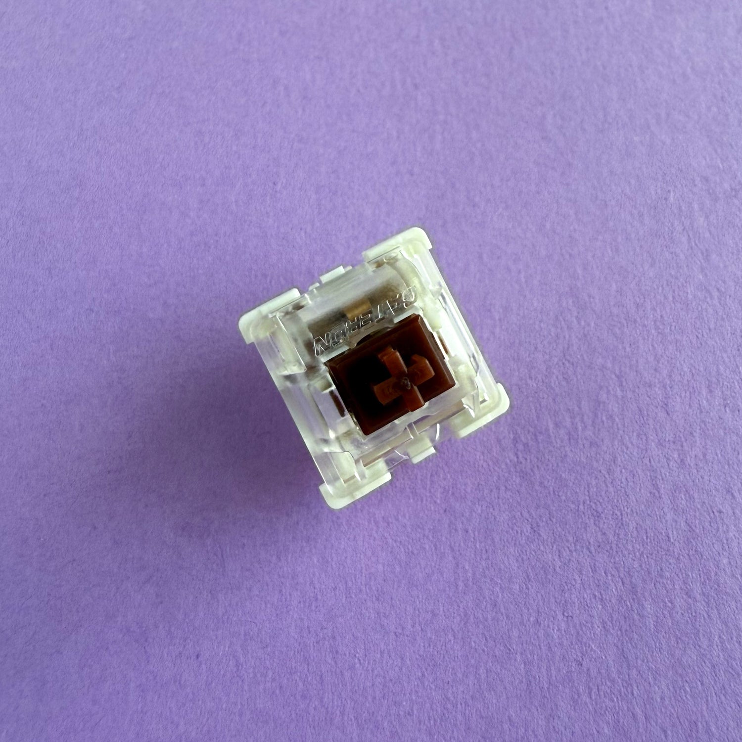 A tactile keyboard switch on a purple background. The switch component is brown, the bottom of the casing is white, and the top of the casing is clear with the word "GATERON" imprinted into it.
