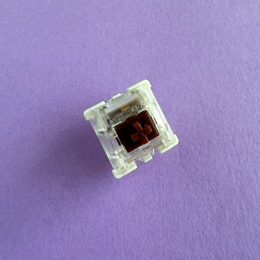A tactile keyboard switch on a purple background. The switch component is brown, the bottom of the casing is white, and the top of the casing is clear with the word "GATERON" imprinted into it.