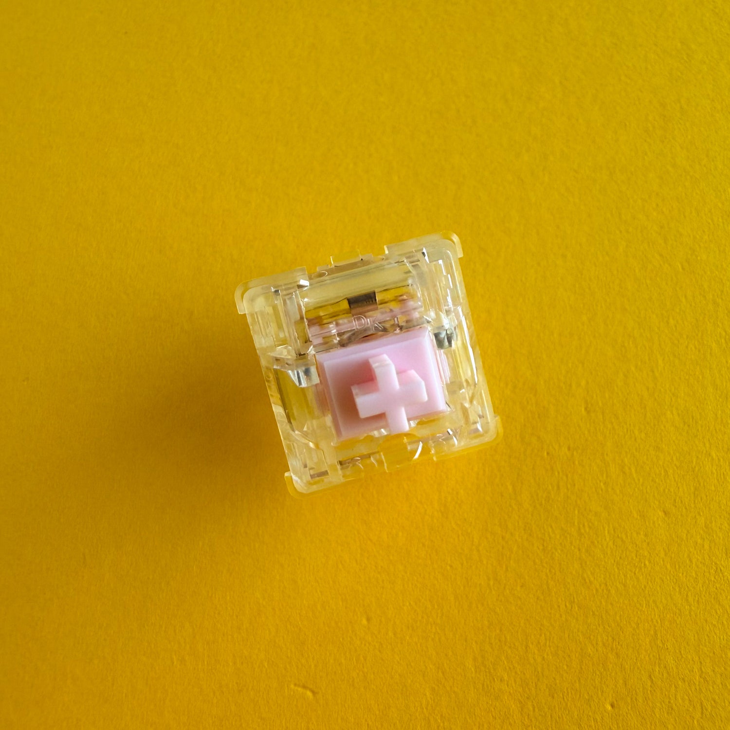 A tactile keyboard switch on a yellow background. The switch component is light pink, the casing is clear. 