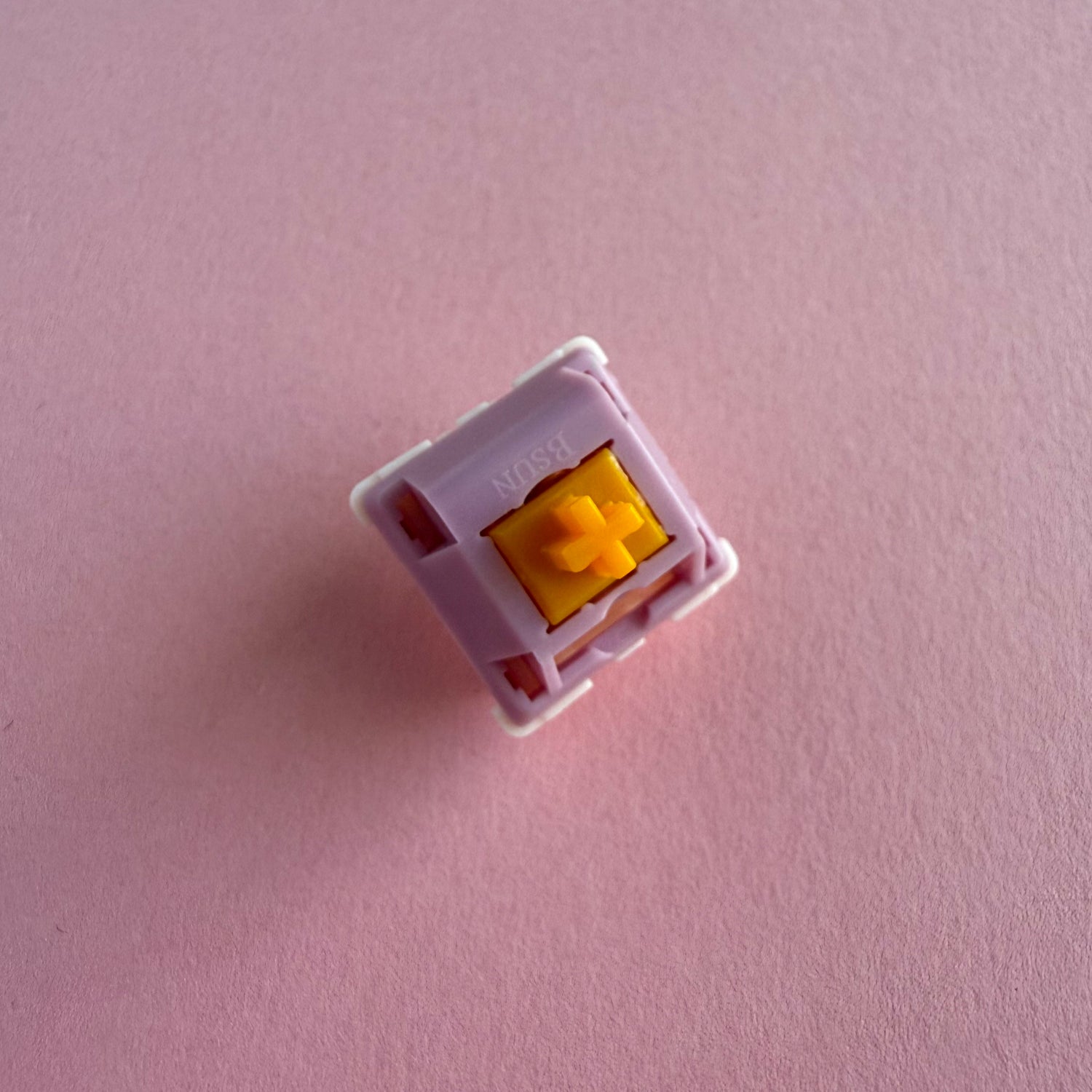 A linear keyboard switch on a purple background. The switch component is orange, the casing is purple.