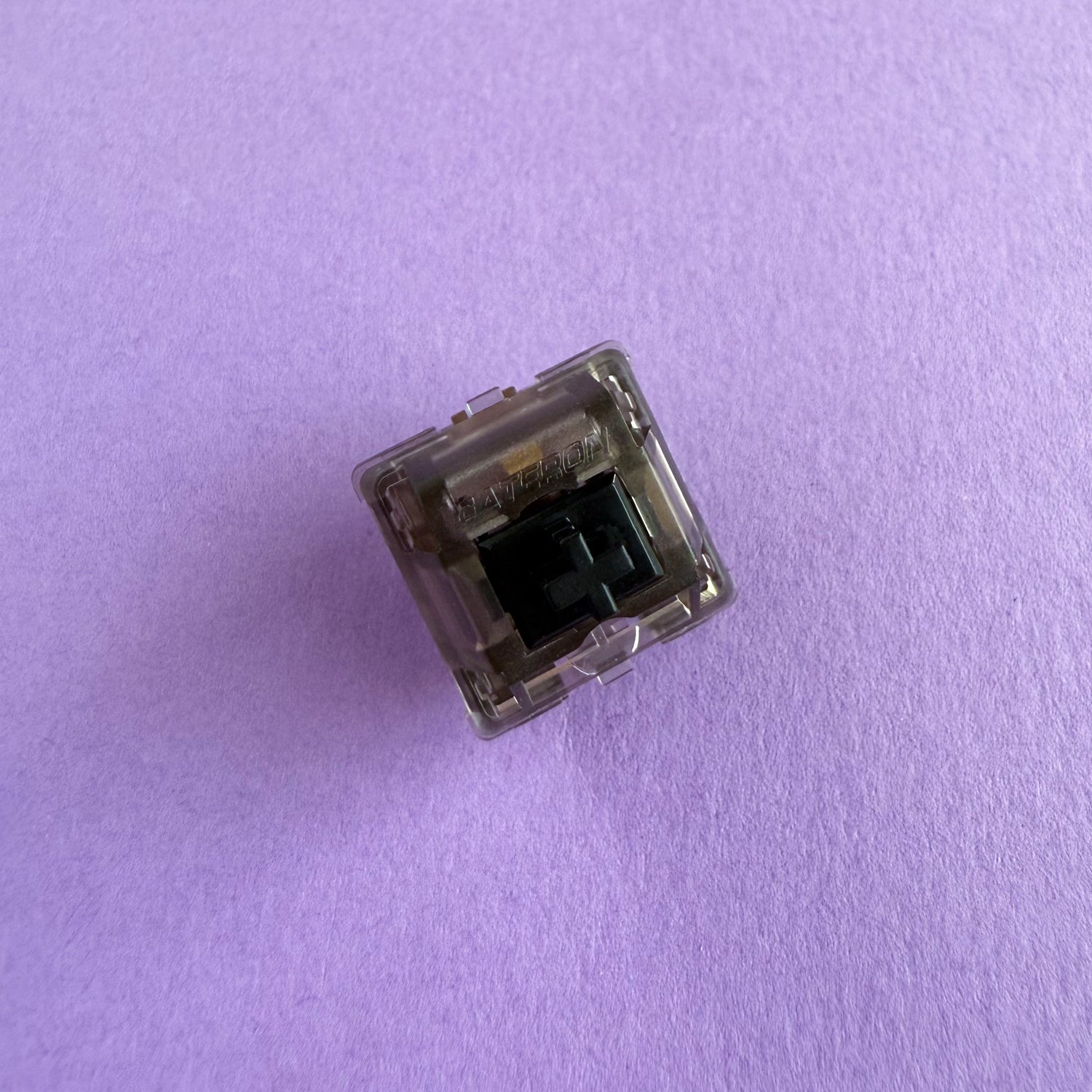 A linear keyboard switch on a purple background. The switch component is black, and the casing is a translucent black with the word "GATERON" imprinted into it.