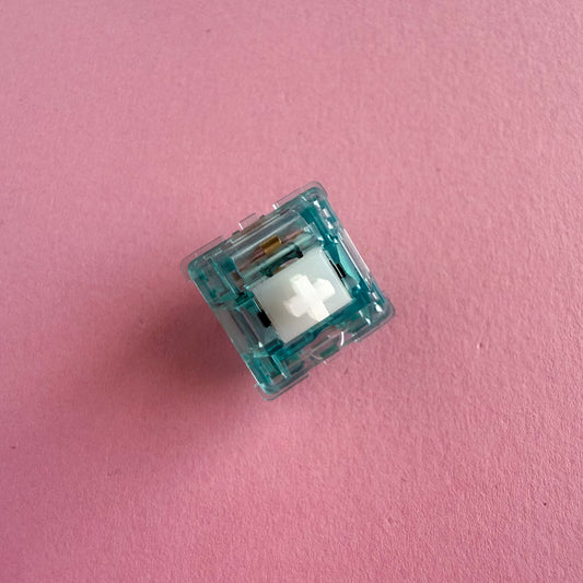 A tactile keyboard switch on a pink background. The switch component is white, and the casing is a translucent blue.