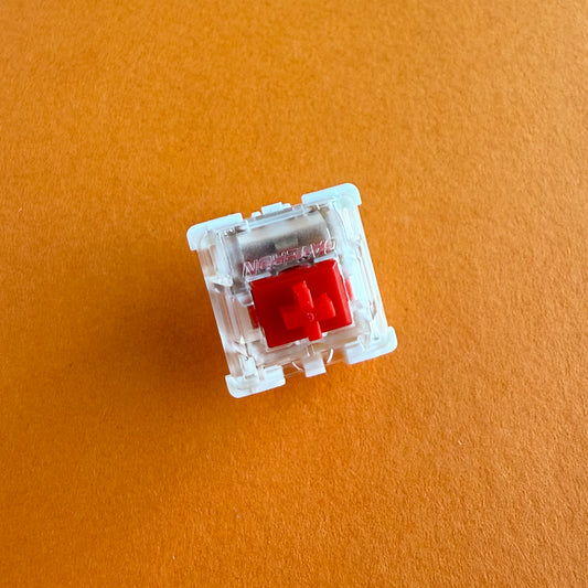A linear keyboard switch on an orange background. The switch component is red, the bottom of the casing is white, and the top of the casing is clear with the word "GATERON" imprinted into it.