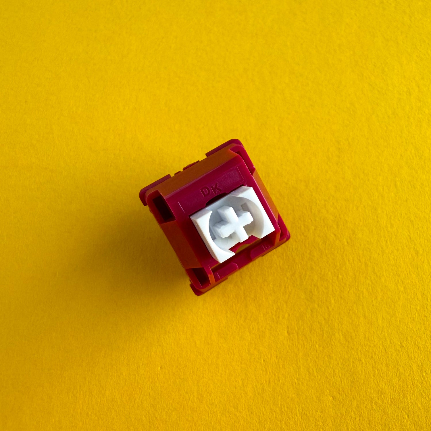 A tactile keyboard switch on a yellow background. The switch component is white, and the casing is a deep red with the letters "DK" imprinted into it.