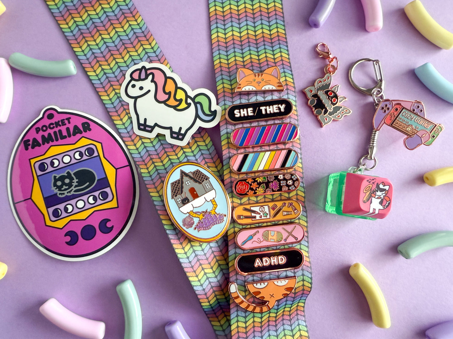 Purple background with pastel colored macaroni shaped beads around. Products are being displayed. From right to left: pink tamagotchi shaped sticker with a black cat, rainbow knit lanyard, mini chubby unicorn sticker, knitting baba yaga pin, orange car head pin, she/they pin in black, bisexual pin, queer pin, subtle floral pin, art personality pin, craft personality pin, adhd pin in black, orange cat butt pin, mothman charm, clicky cat with the jazz cat design, and cozy gamer pin.