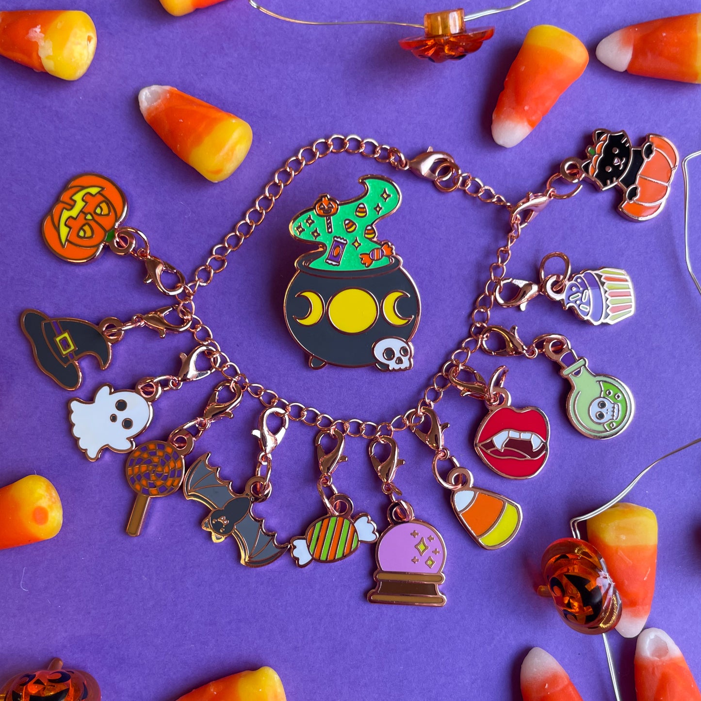 A charm bracelet with 12 Halloween themed charms on it and an enamel pin of a cauldron in the center of the bracelet.