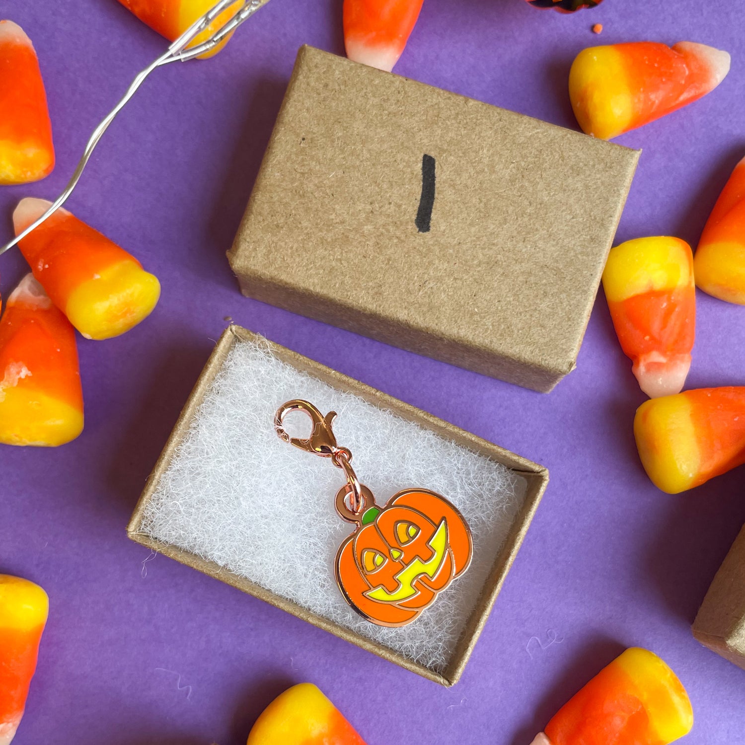 A Kraft paper box with a 1 on it the box contains a charm with a lobster claw clasp shaped like a jack-o-lantern. The box is on purple paper scattered with candy corn.