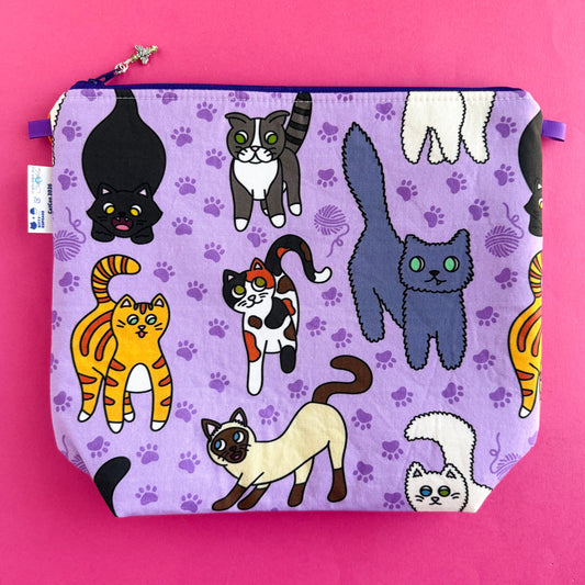 Product photo of bag collaboration between Kitty With A Cupcake and Stitched by JessaLu. The zipper has a signature SbJL charm. This photo is showing the front of the bag which is cat illustrations by KWAC. There is a black cat, tabby, orange cat, calico, grey cat, siamese, and white cat visible. The cats are on a purple background with darker purple paw prints and yarn balls filling in the negative space. All on a hot pink background.