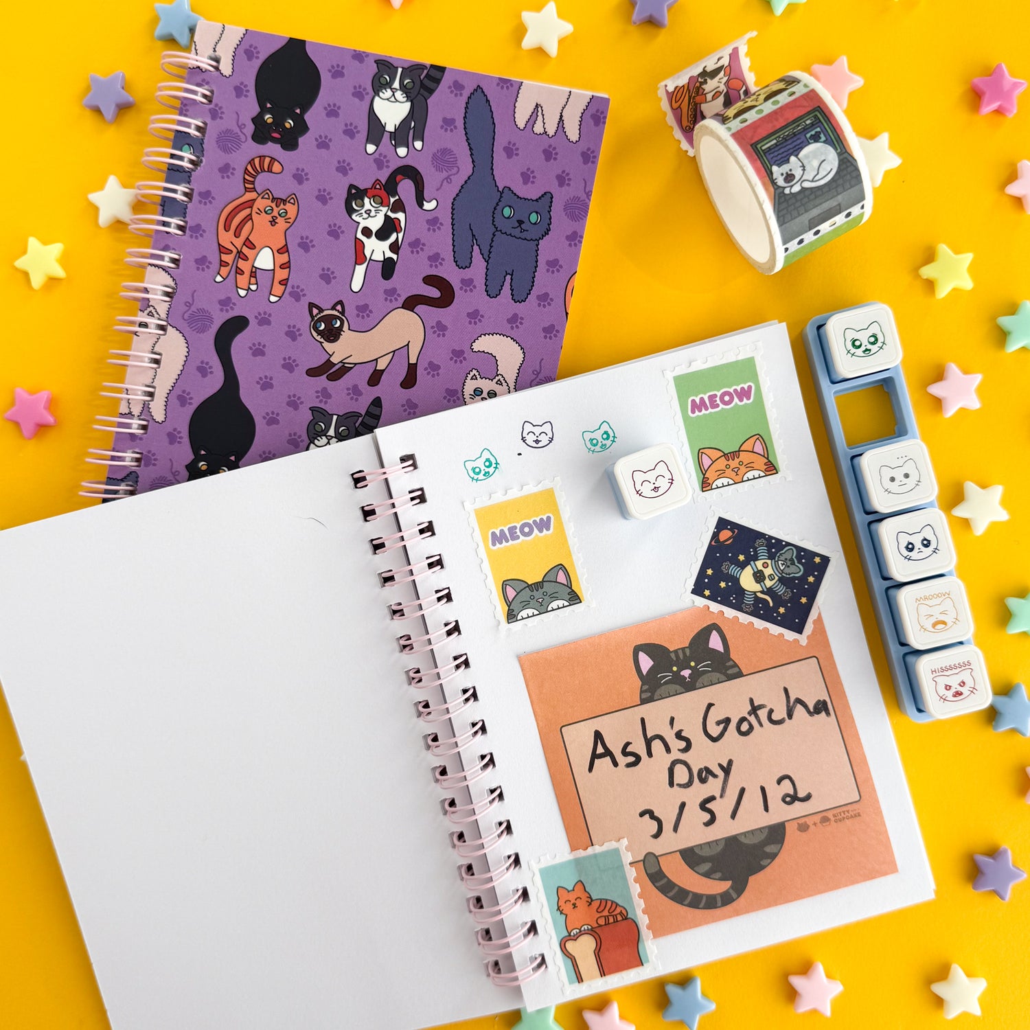 An open notebook with a spread of cat themed stationery on it like cat washi tape stamps, push button cat stamps, and a cat sticky note that reads "Ash's Gotcha Day 3/5/12". The open notebook is lying on a notebook with a purple cover with cat illustrations on it.