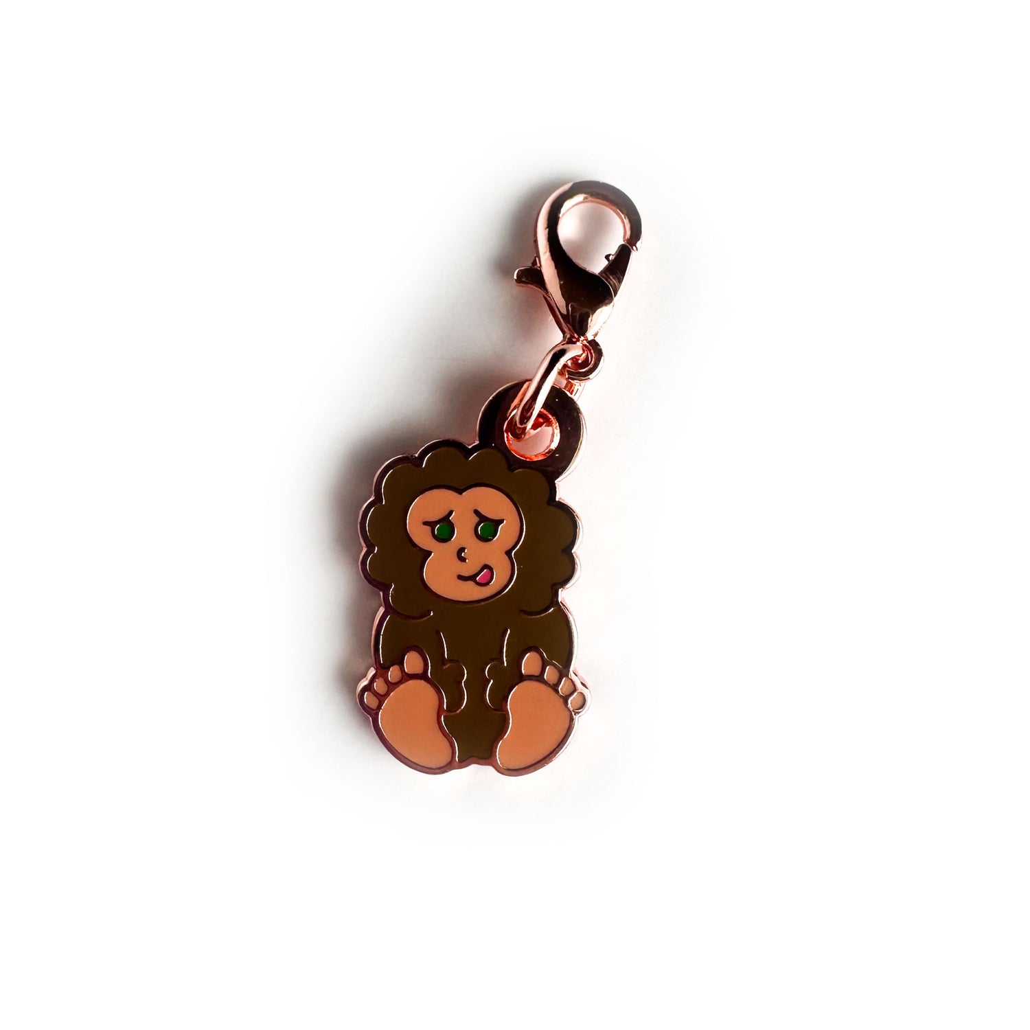 An enamel charm in the shape of a cartoon Bigfoot sitting down with a cute sheepish smile, the charm has a lobster claw clasp. 