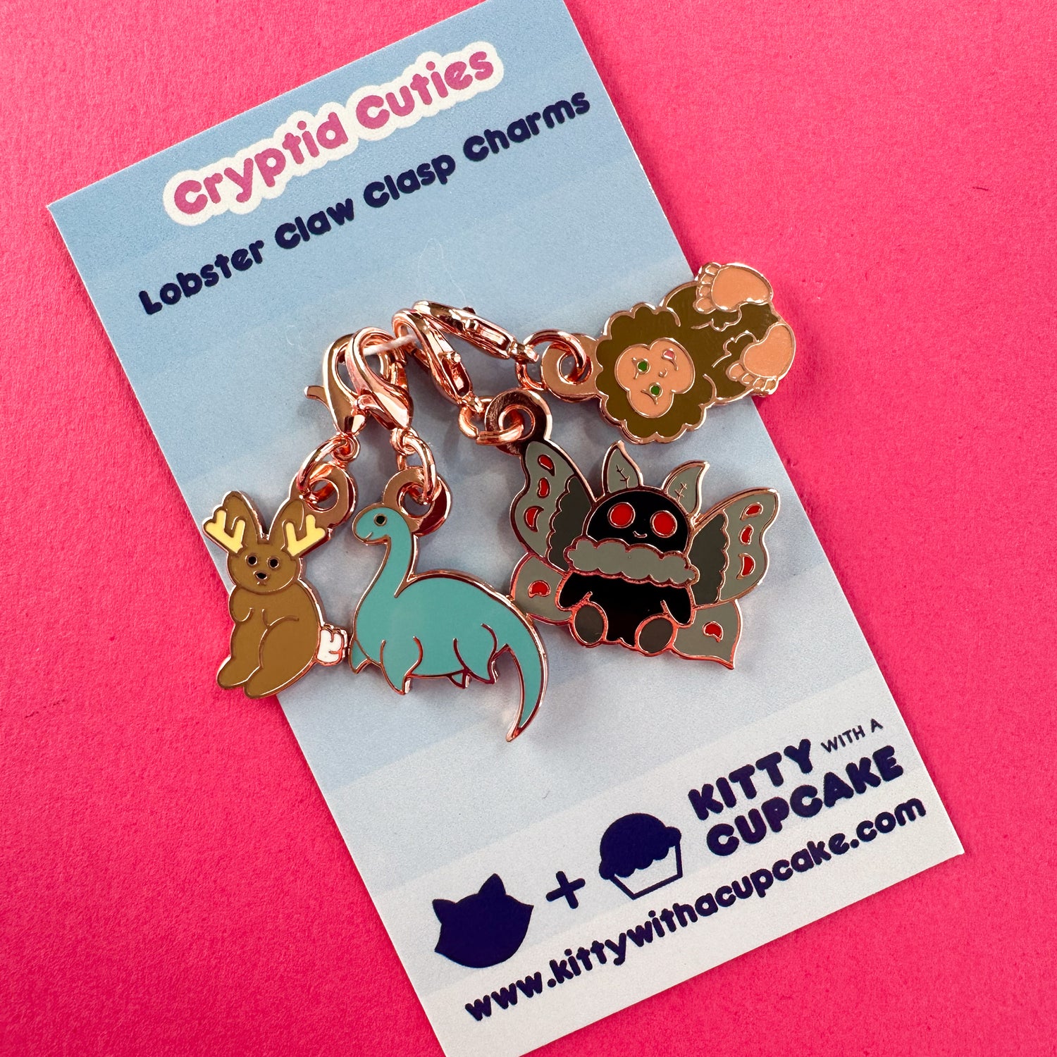 A card with 4 lobster claw clasp charms attached to it. The card reads "Cryptid Cuties - Lobster Claw Clasp Charms". The charms are shaped like a jackalope, Loch Ness Monster, Mothman, and Bigfoot. 