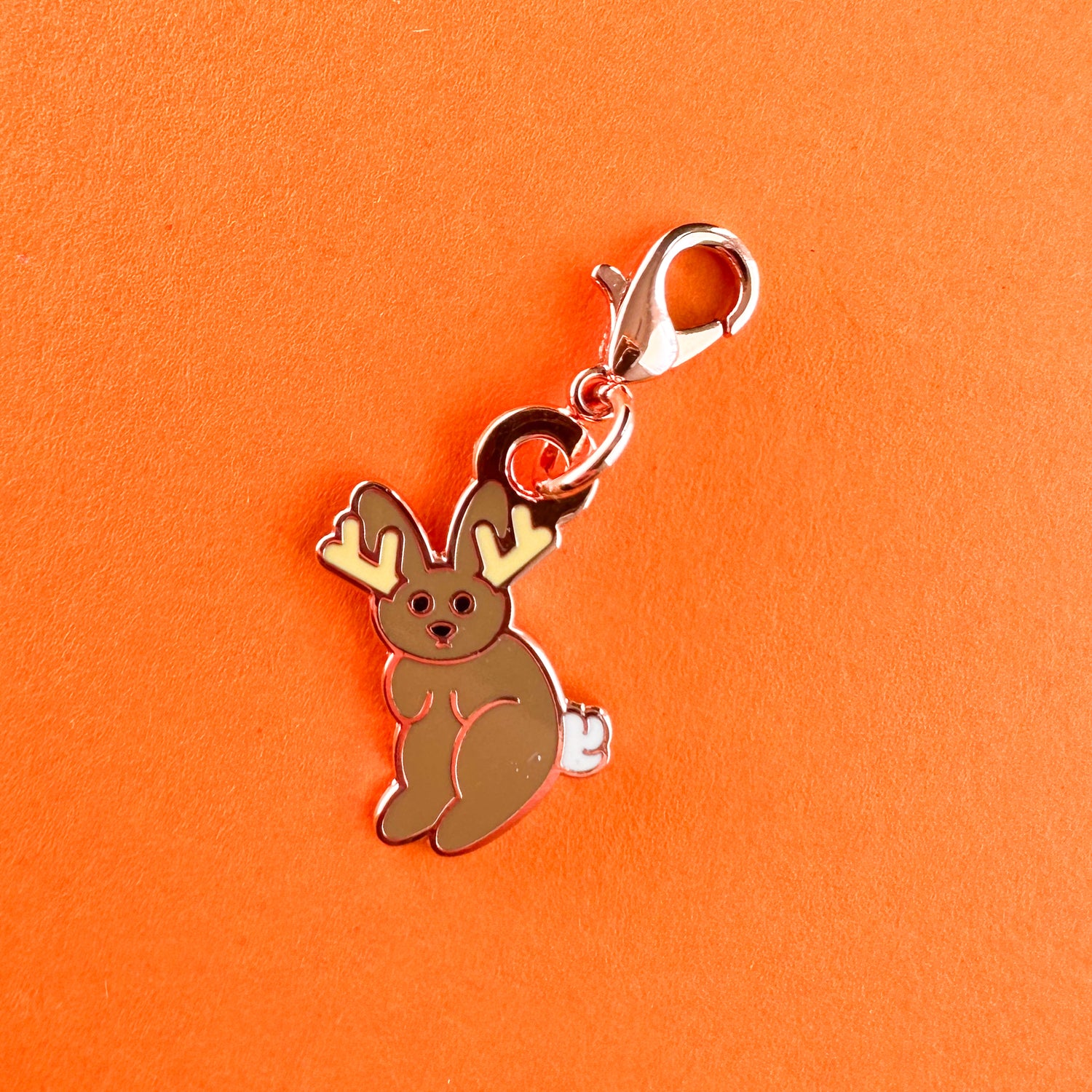 An enamel charm in the shape of a cute cartoon style illustration of a jackalope, a rabbit with antlers. 