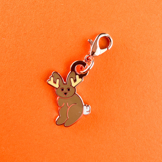 An enamel charm in the shape of a cute cartoon style illustration of a jackalope, a rabbit with antlers. 