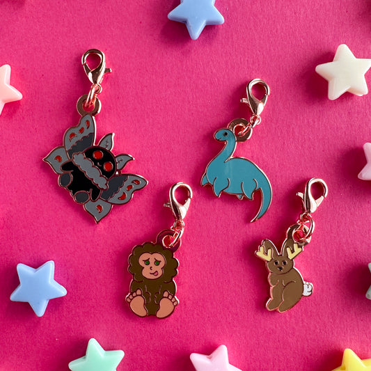 Four charms in the shape of cute cryptids including Mothman, Bigfoot, Nessie, and a jackalope. The charms all have lobster claw clasps. The charms are on a pink paper background with plastic pony beads around them.