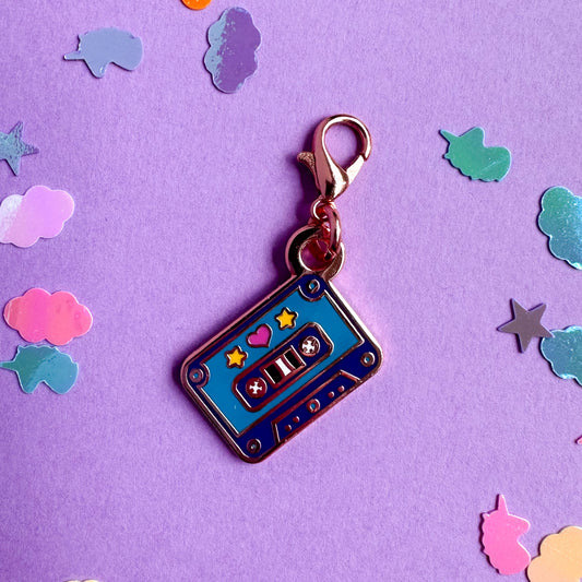 A lobster claw clasp charm of a purple and teal cassette tape on a purple confetti background