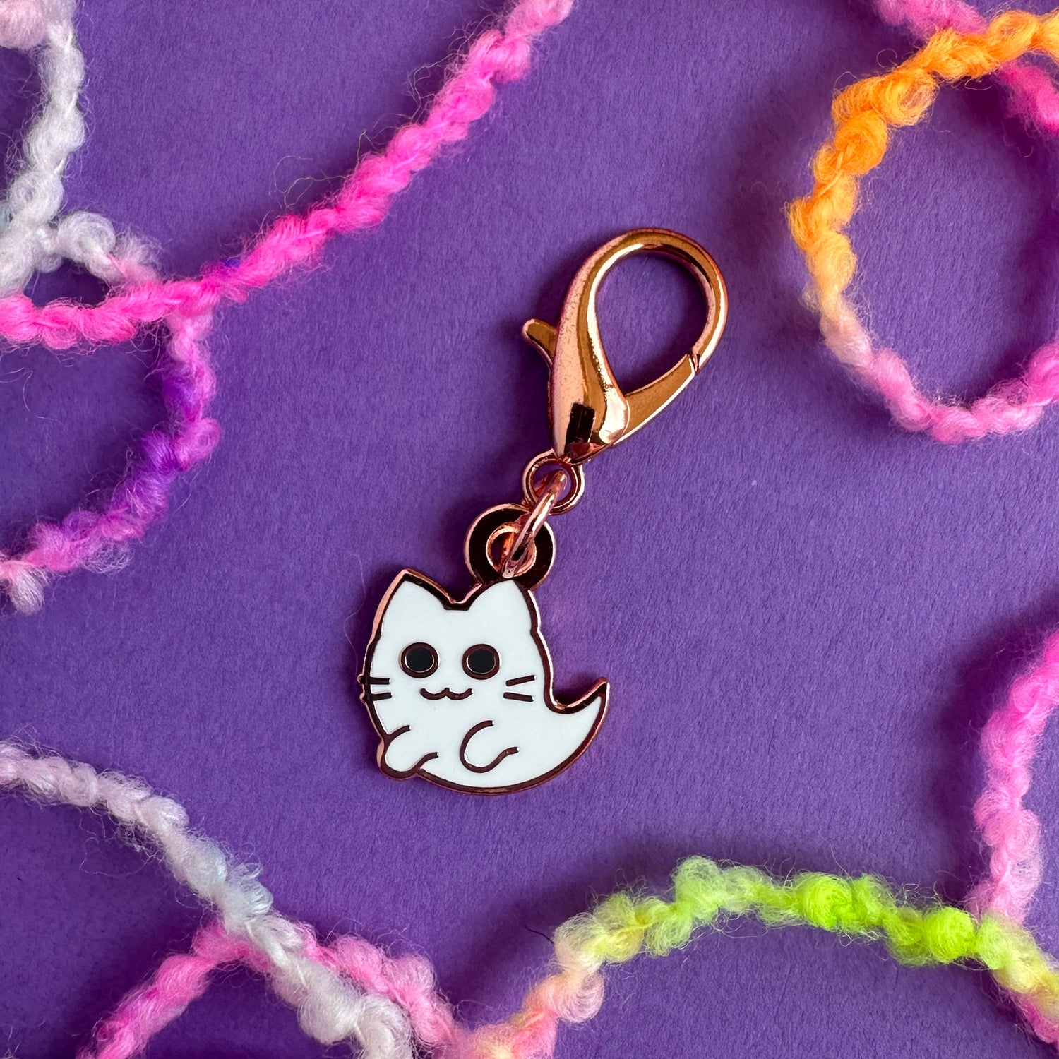 A cute ghost kitty charm with a lobster claw clasp on a purple background surrounded by brightly colored yarn. 