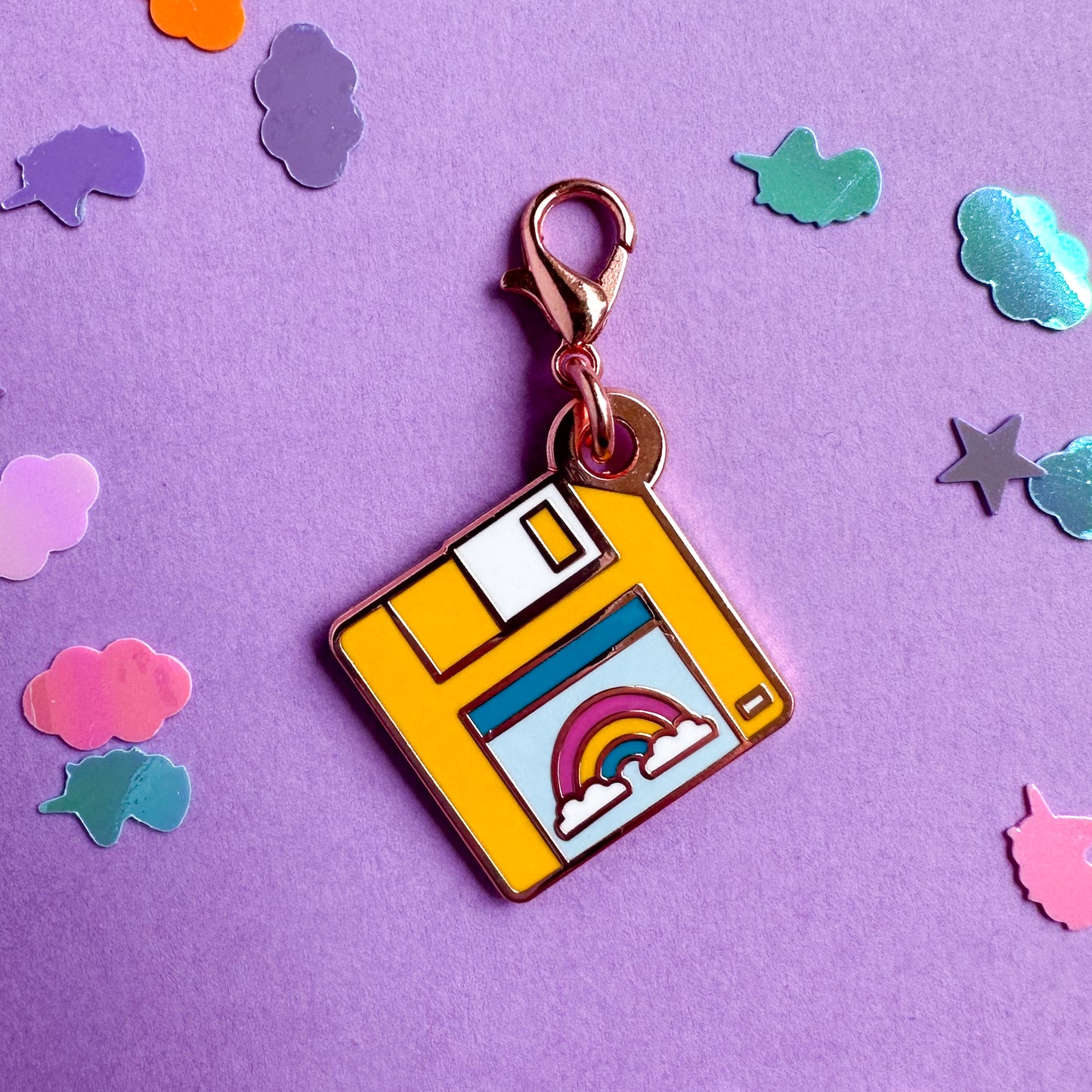 A lobster claw clasp charm shaped like a yellow floppy disk with a rainbow on the label. The charm is on a lavender confetti background.