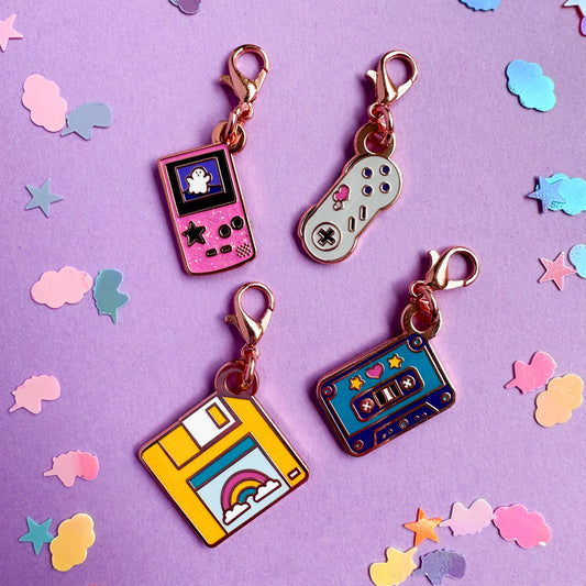 A group of charms shaped like a gameboy color, Super Nintendo controller, floppy disk, and cassette tape on a lavender confetti background.