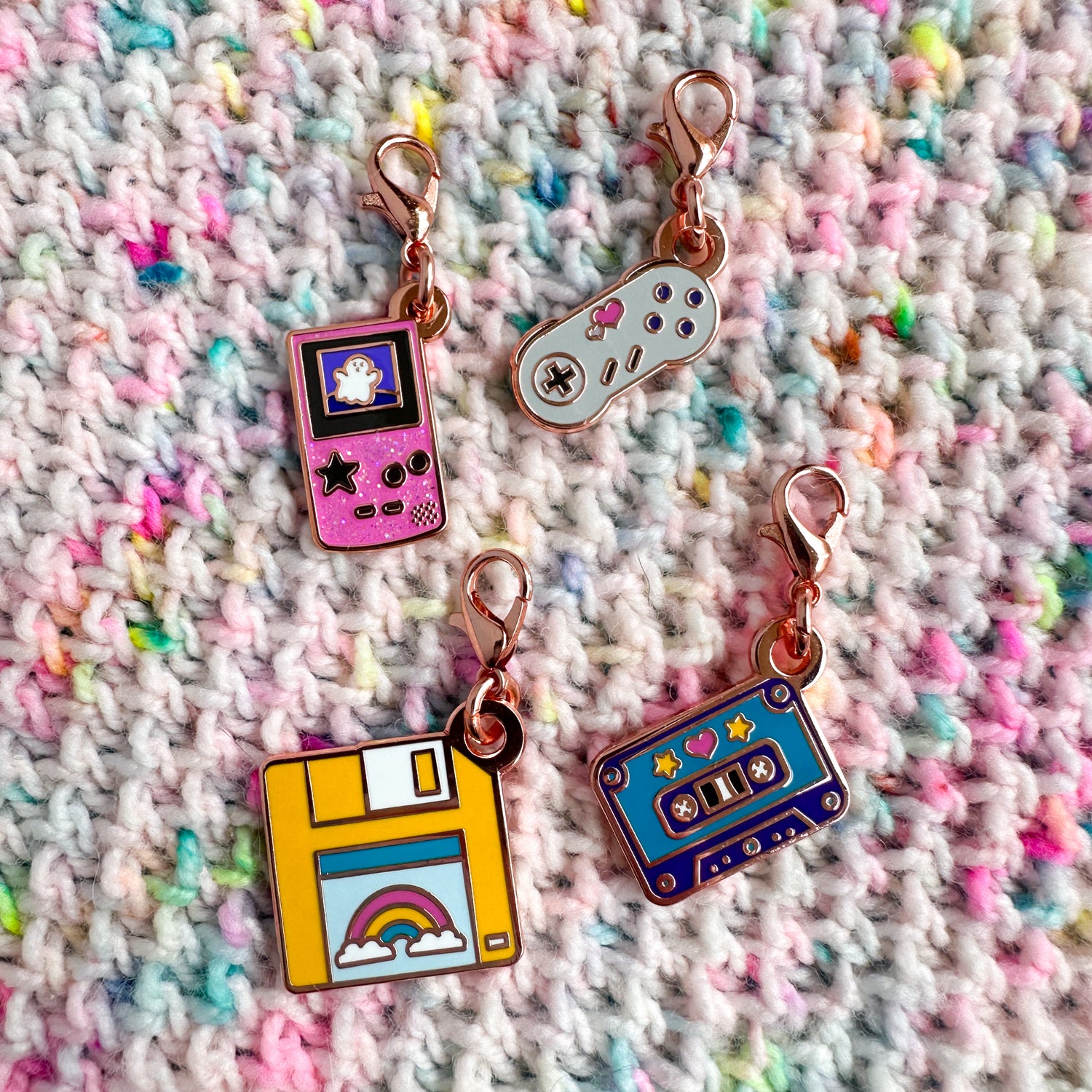A set of four lobster claw clasp charms on a brioche stitch knitted background in pastel confetti yarn. The charms are shaped like a gameboy, game controller, floppy disk, and cassette tape.