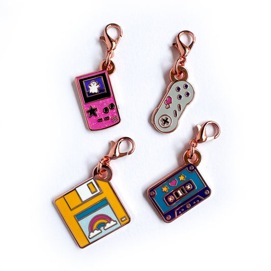 A set of lobster claw clasp charms, one is shaped like a pink gameboy with a ghost on it, another is shaped like a Super Nintendo controller with hearts, a third is shaped like a yellow floppy disk with a rainbow on it, and the 4th is shaped like a purple cassette tape. 