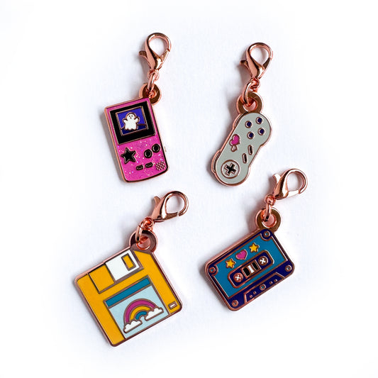 A set of lobster claw clasp charms, one is shaped like a pink gameboy with a ghost on it, another is shaped like a Super Nintendo controller with hearts, a third is shaped like a yellow floppy disk with a rainbow on it, and the 4th is shaped like a purple cassette tape.