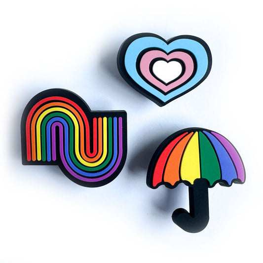 Three PVC charms one in the shape of a heart in the colors of the Trans Pride flag, one a rainbow squiggle, and one a rainbow umbrella.