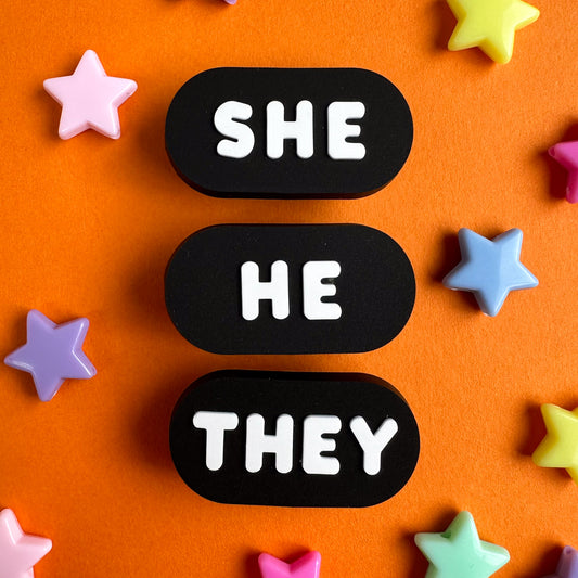 Three black oval shoe charms with different pronouns written on them in bubble letters, one reads "She", one reads "He", and one reads "They" the charms are resting on an orange piece of paper with star beads around them. 