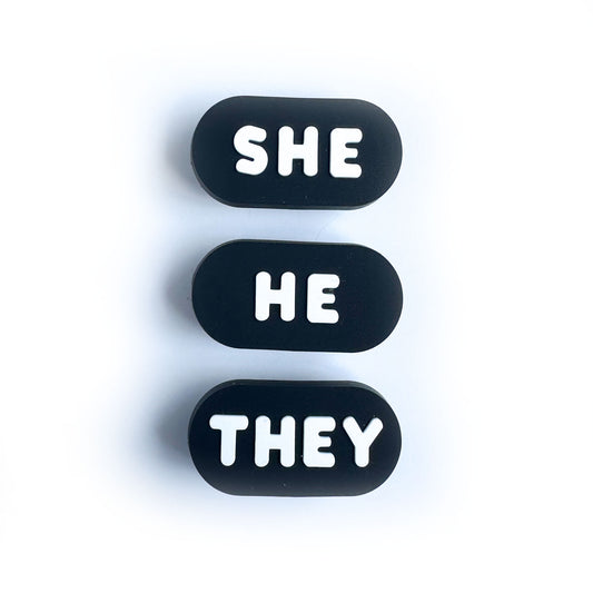 Three black oval shaped shoe charms each with white bubble letters, one says "She", another "He", and the last one "They"