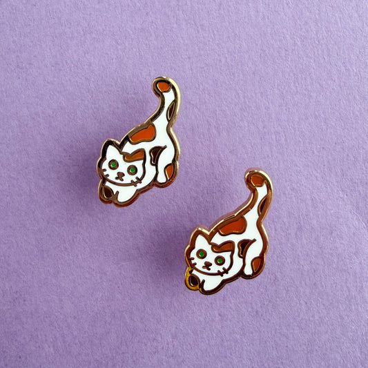 Product photo of the Calico Cat Earrings. This cat has green eyes and a white body with orange and brown spots. There are two earrings displayed diagonal from each other. The earrings are resting against a purple background.