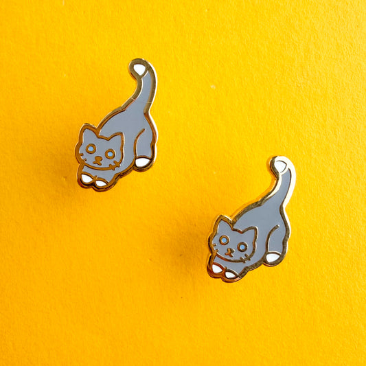 Product photo of the Grey Cat Earrings. This cat has blue eyes and a grey body with a white tail tip and feet. There are two earrings displayed diagonal from each other. The earrings are resting against a yellow background.