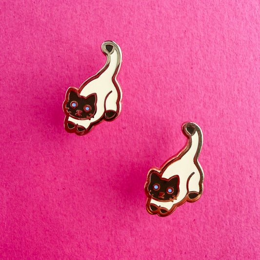 Product photo of the Siamese Cat Earrings. This cat has blue eyes and a cream body with a brown head, tail tip, and feet. There are two earrings displayed diagonal from each other. The earrings are resting against a hot pink background.