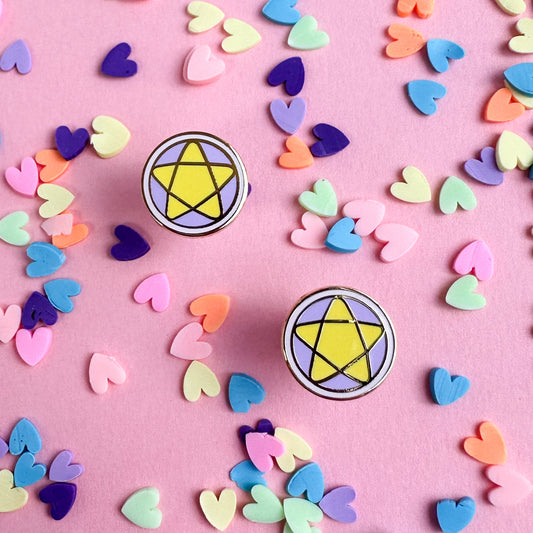 Stud earrings in the shape of pentacles with pastel pink and lavender coloring and yellow stars on top of a pink paper background with pastel heart confetti on it.