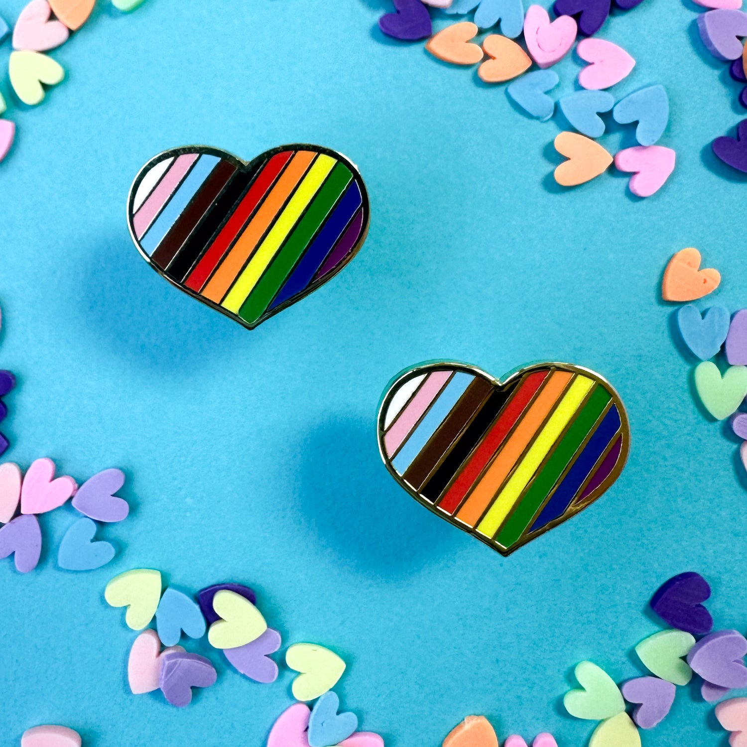 Two earrings in the shape of chunky hearts with diagonal stripes of the inclusive pride flag. The earrings are on a blue background with pastel heart confetti around. 