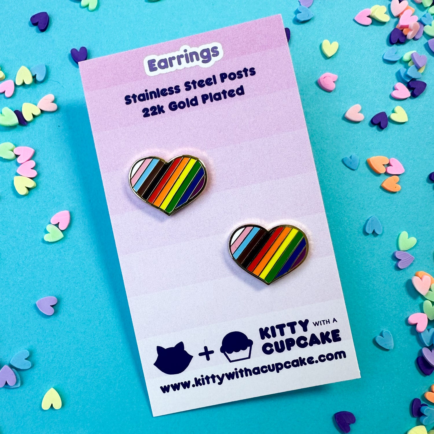Two chunky heart earrings with diagonal stripes on them in the progress pride flag colors. The earrings are packaged on a pink card that is sitting on top of a blue background with confetti hearts around it. 