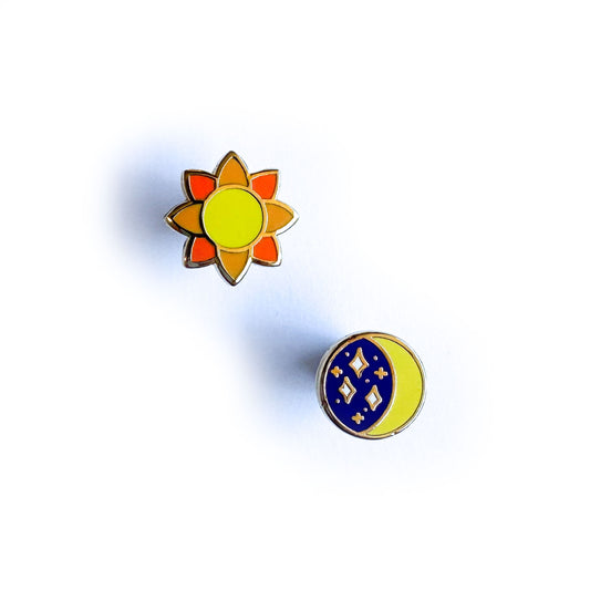 A set of earrings on a white background. One earring is sun shaped with a yellow circle center, and petals of two shades of orange. The other is a circle, with a yellow crescent moon and the rest of the circle is blue with stars.
