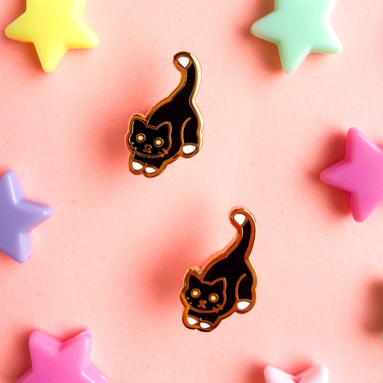 Tuxedo cat shaped post earrings with gold details with colorful star beads around them on a pink background. 