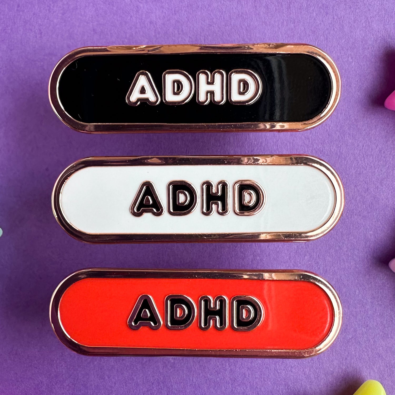 Three capsule shaped pins that have the letters ADHD on them, the top is black, the middle is white, and the bottom is red.  The pins are on a purple paper background. 