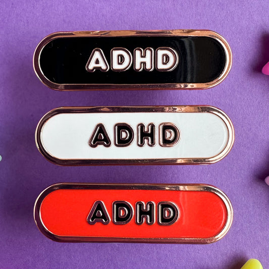 Three capsule shaped pins that have the letters ADHD on them, the top is black, the middle is white, and the bottom is red.  The pins are on a purple paper background. 