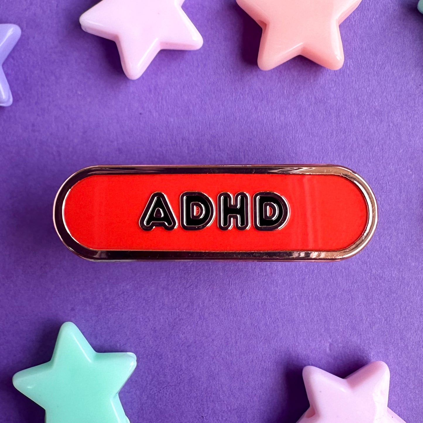 A bandaid shaped enamel pin with black bubble letters on it that read "ADHD" on a red background. The pin is sitting on a purple piece of paper with pastel star beads around it. 