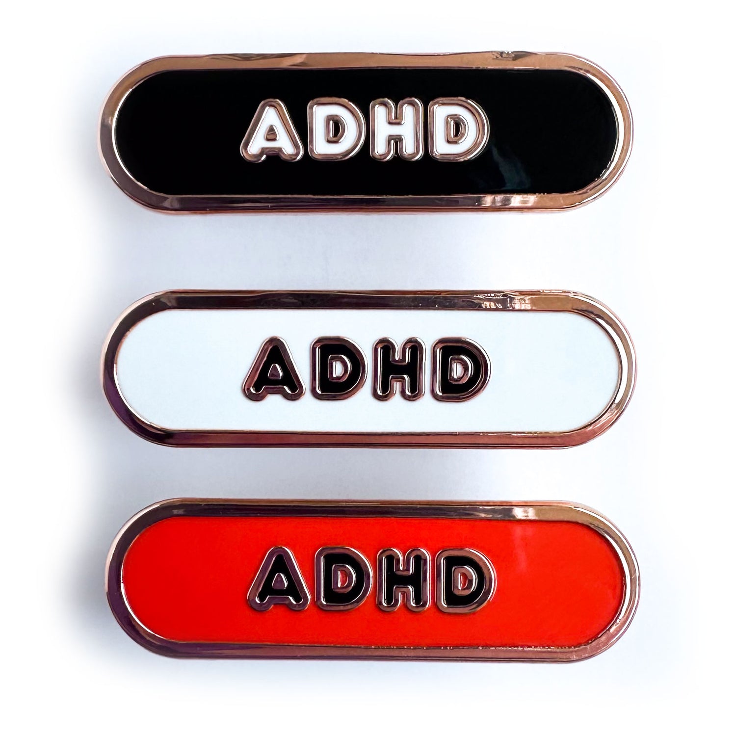 Three capsule shaped pins that have the letters ADHD on them, the top is black, the middle is white, and the bottom is red. 