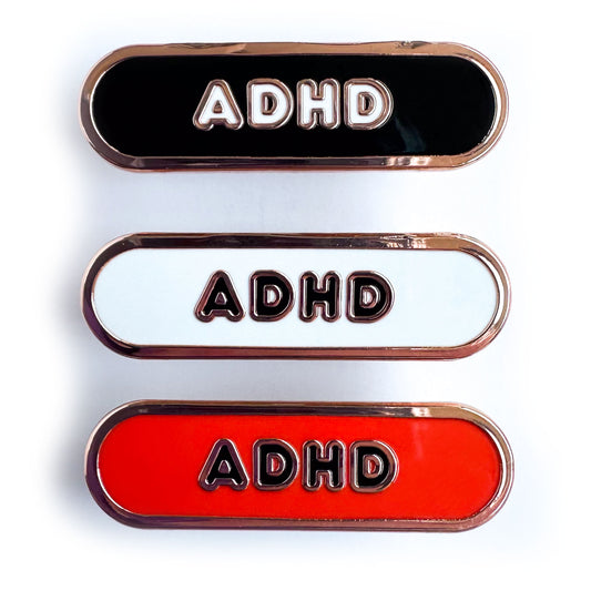 Three capsule shaped pins that have the letters ADHD on them, the top is black, the middle is white, and the bottom is red.