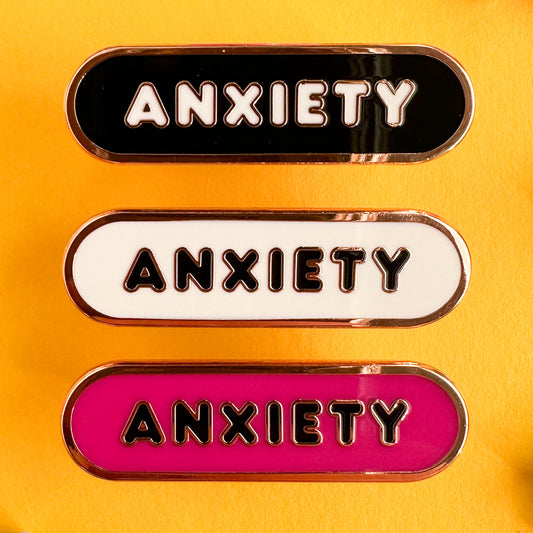 A set of three enamel pins all with bubble letters that spell "Anxiety" on them, the top one is black, the middle is white and the bottom is hot pink. All of the pins are on a yellow background.