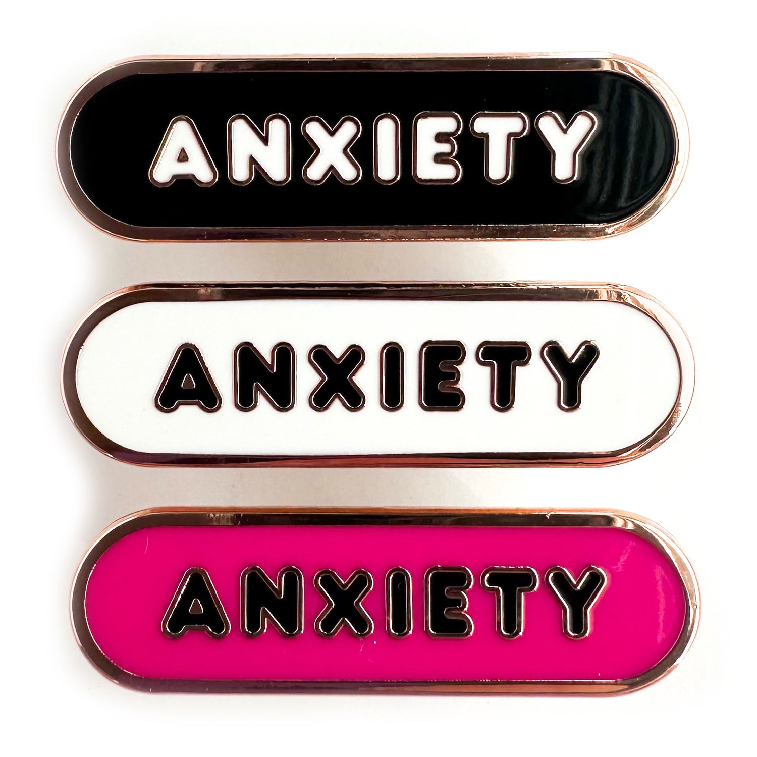 A set of three enamel pins all with bubble letters that spell "Anxiety" on them, the top one is black, the middle is white and the bottom is hot pink.