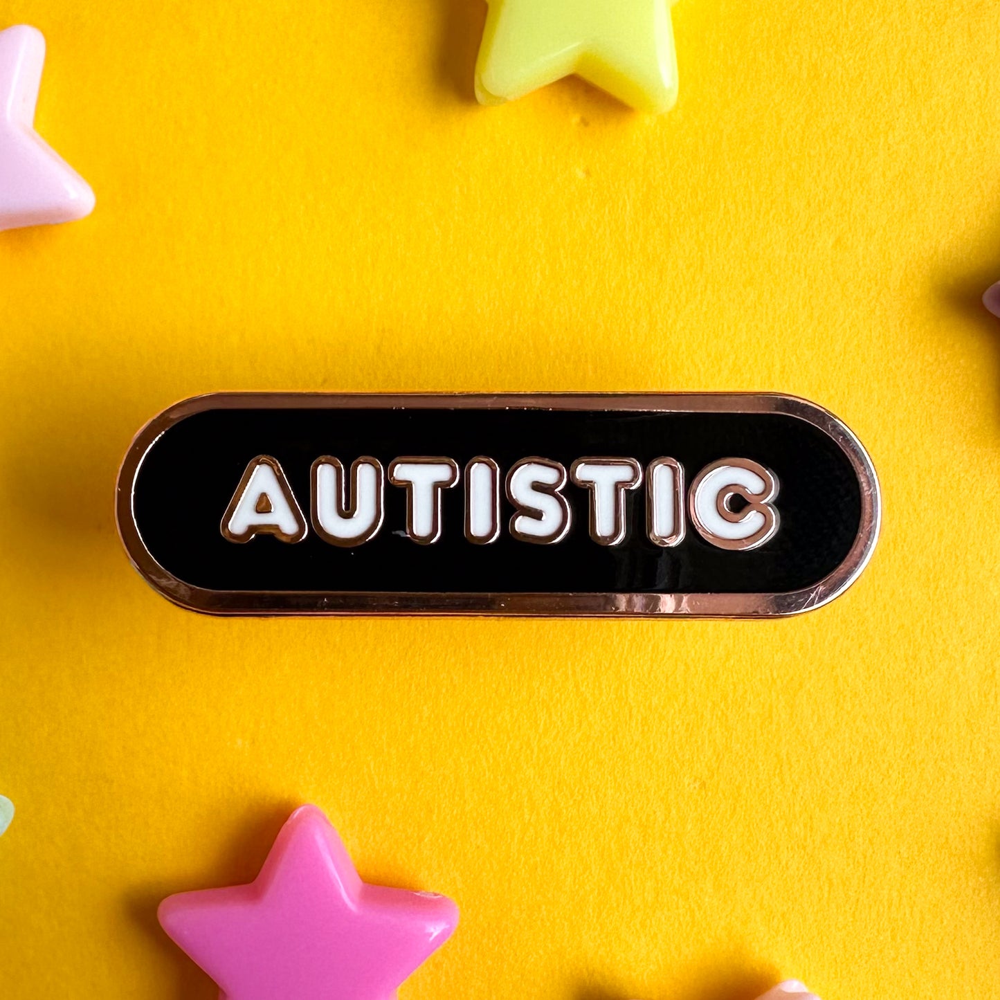 A black oval pin with white bubble letters that read "Autistic". The pin is sitting on a yellow paper background with pastel star beads around it. 