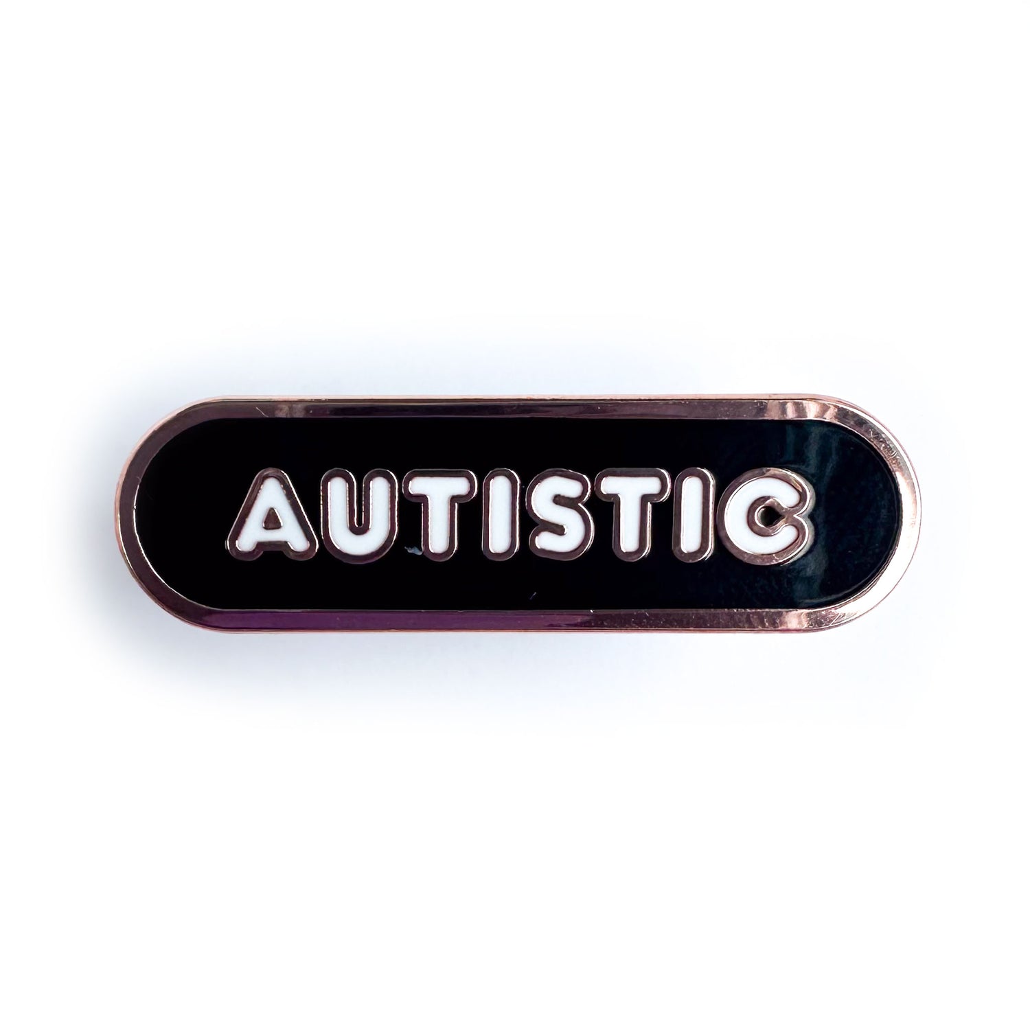 An enamel pin with rose gold colored metal that is in the shape of a black capsule with white bubble letters that spell "Autistic".