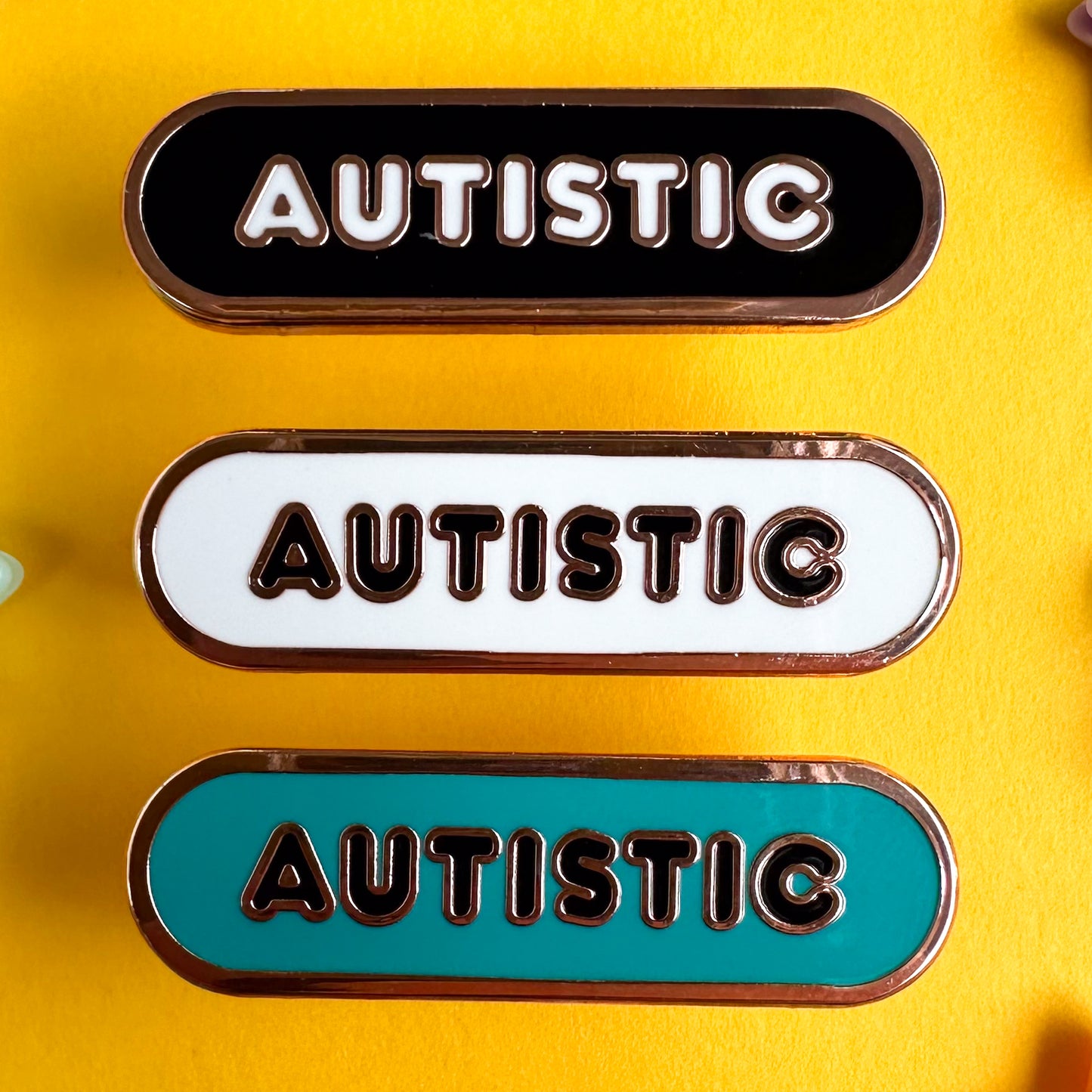 Three enamel pins that are all capsule shaped with bubble letters that read "Autistic" on them. The top one is black, the middle is white, and the bottom is teal. The pins are on a yellow paper background.