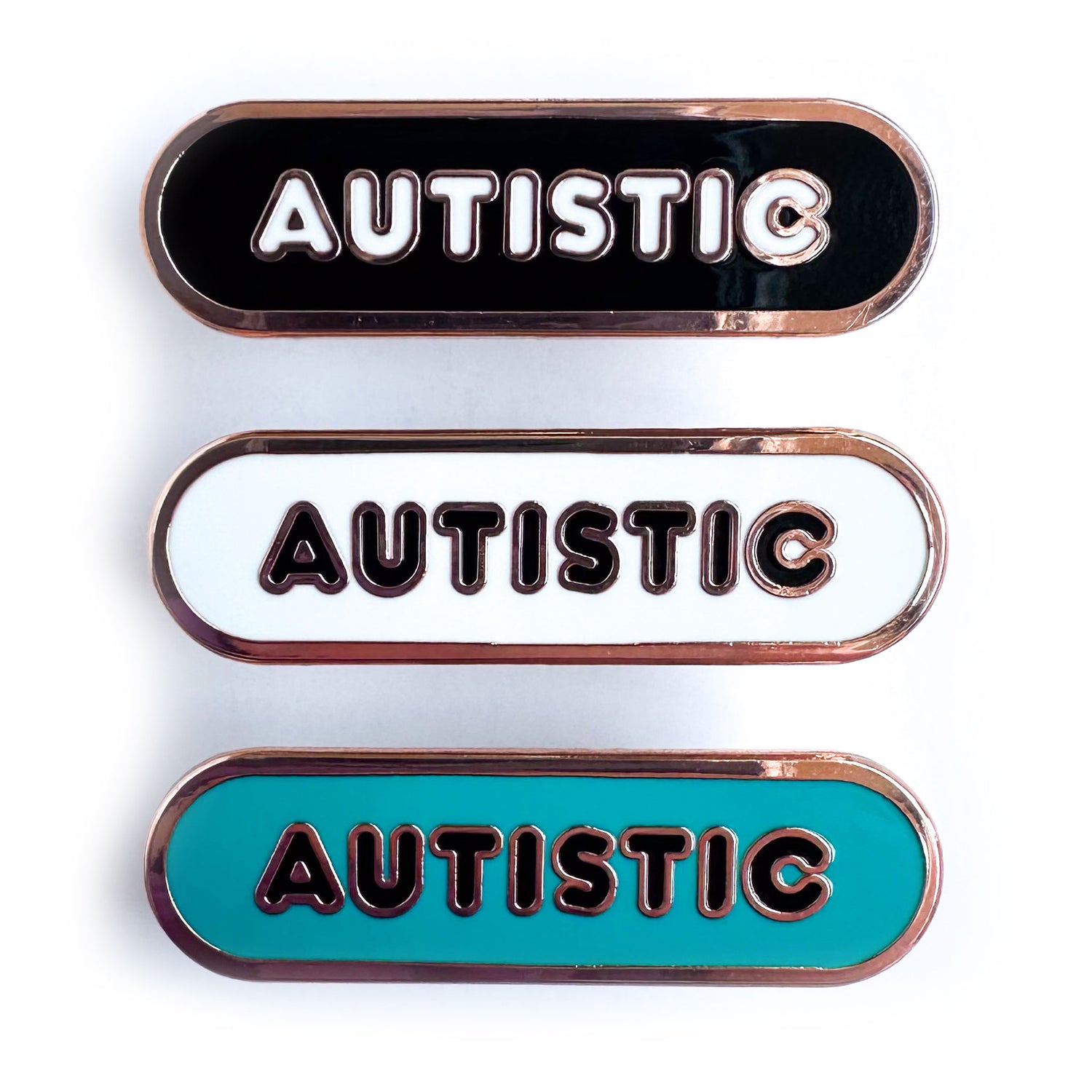 Three enamel pins that are all capsule shaped with bubble letters that read "Autistic" on them. The top one is black, the middle is white, and the bottom is teal.